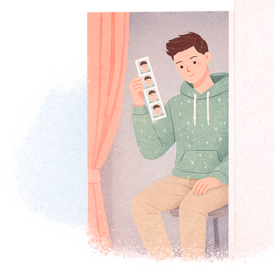 A young man with a dark undercut in a sage hoodie sitting in a photo booth with a coral curtain, holding up a strip of four photos